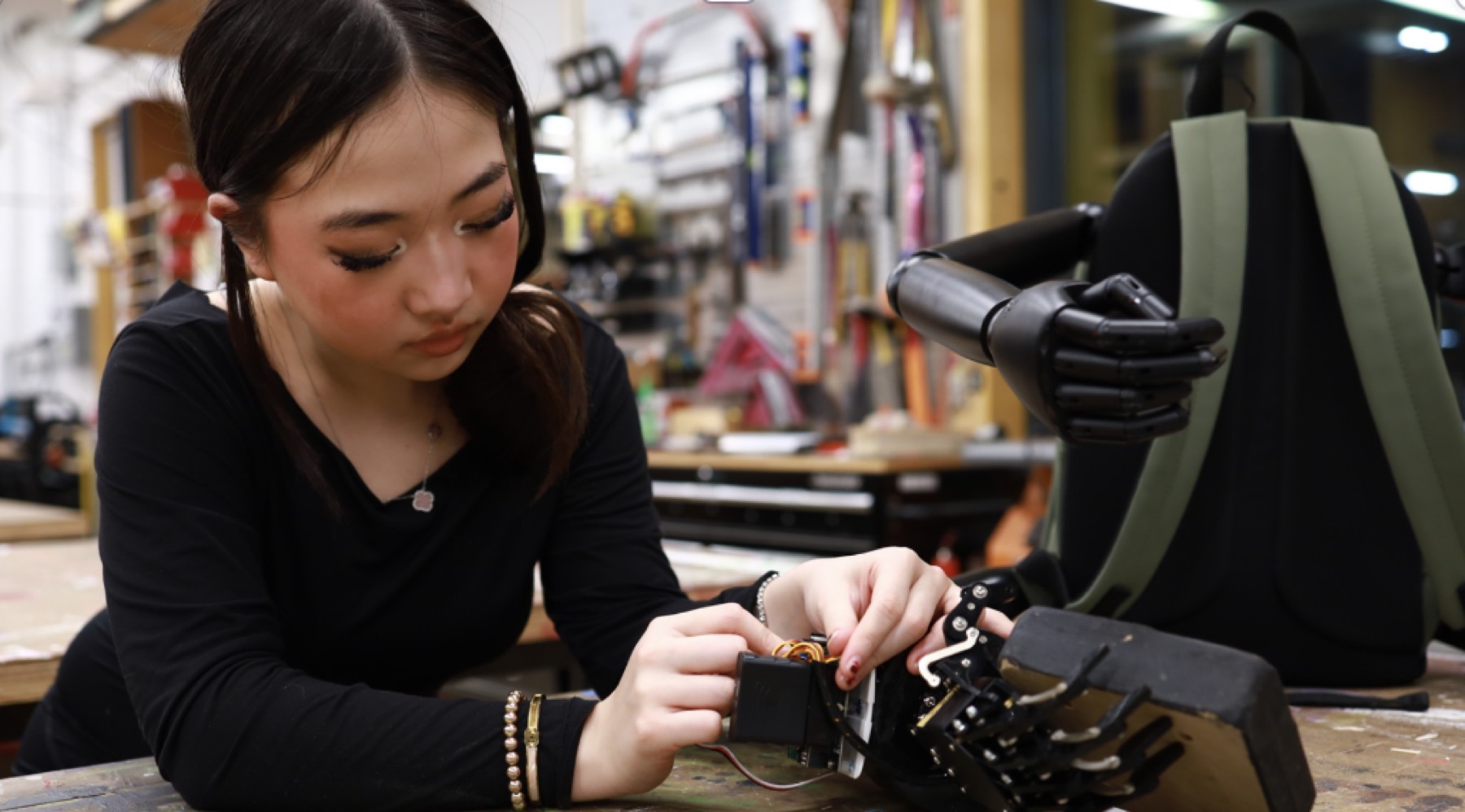 Veronika Wang wiring the electronics of a robotic arm at a workbench, with the ReachOne harness beside her